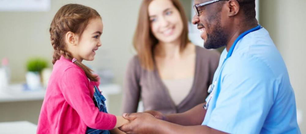 Criança em consulta médica, com a mãe observando ao fundo