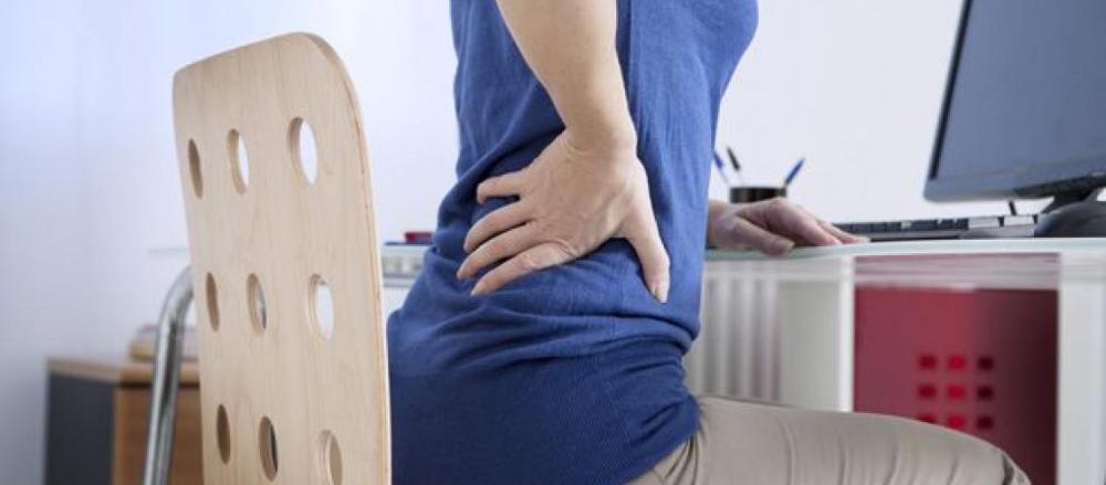 pessoa sentada de frente a um computado com a mão na coluna, indicando dor