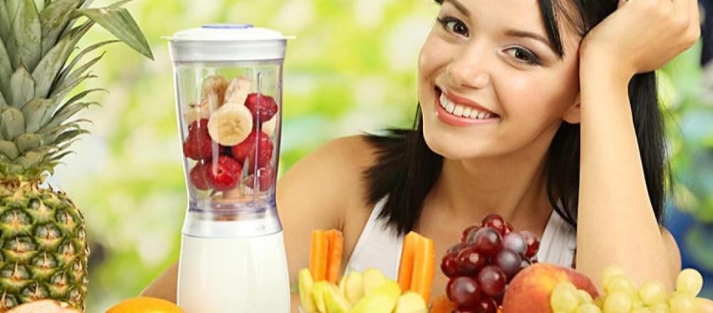 Mulher fazendo uma vitamina em um mini liquidificador em frente a uma mesa cheia de frutas