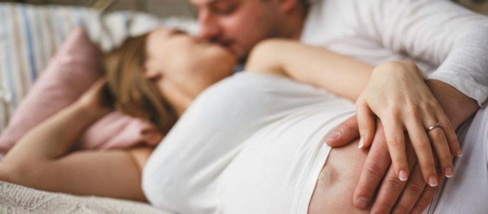 Casal trocando afeto na cama, enquanto seguram a barriga da gestante