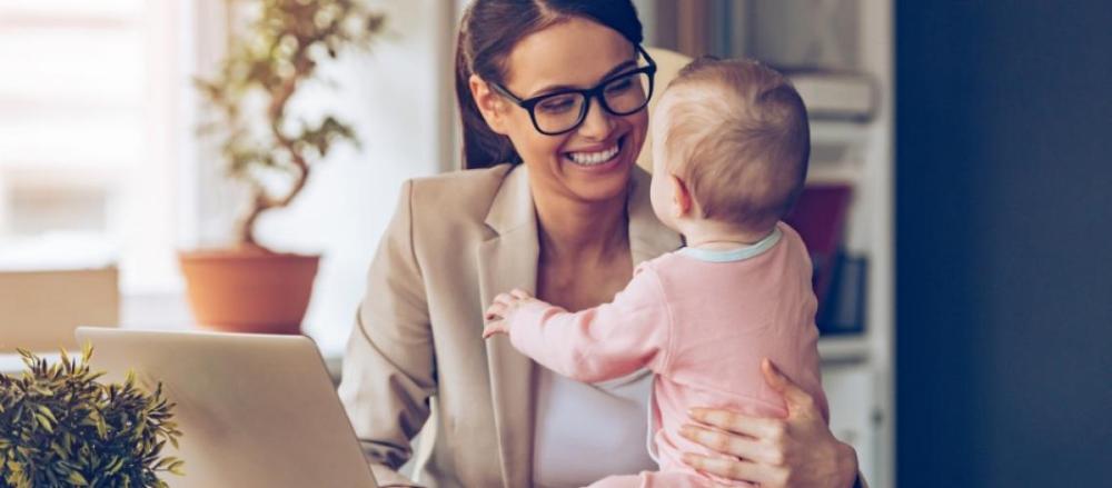 Mulher sorrindo de frente a um computador enquanto segura um bebê