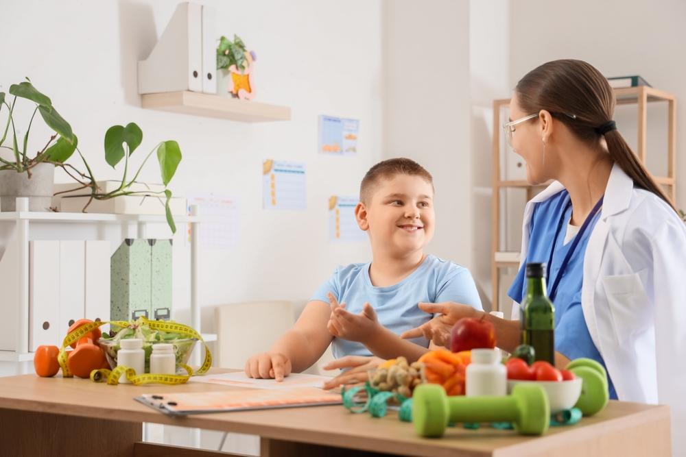 Criança conversando com profissional de saúde em mesa com alimentos saudáveis, pesos e fitas métricas, representando orientação nutricional infantil, microbiota intestinal e prevenção da obesidade infantil.