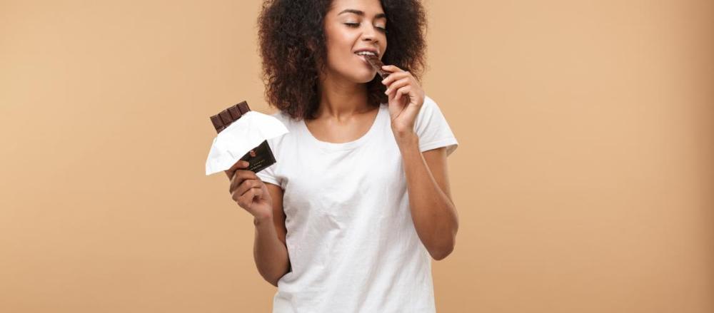 Mulher comendo um pedaço de chocolate enquanto segura uma barra na outra mão