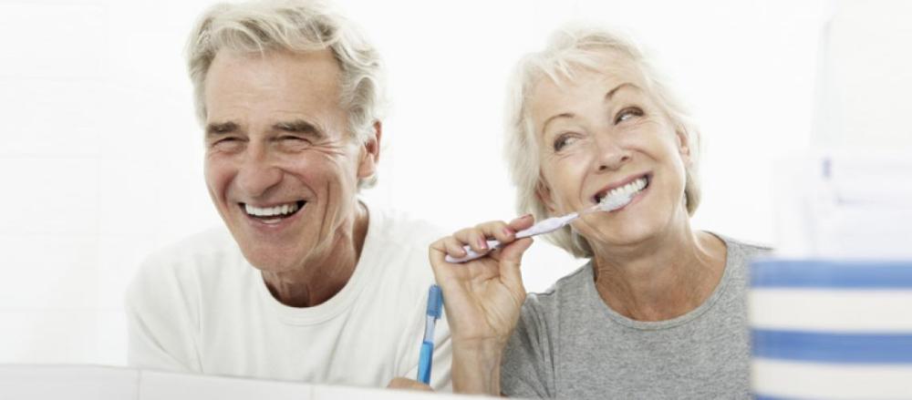 Imagem de um casal idoso em frente ao espelho, com o home sorrindo e amulher escovando os dentes