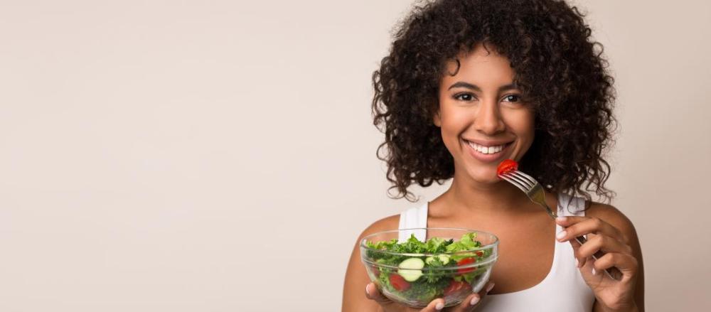 Mulher com um pote de salada na mão, enquanto leva aprte dela a boca com um garfo