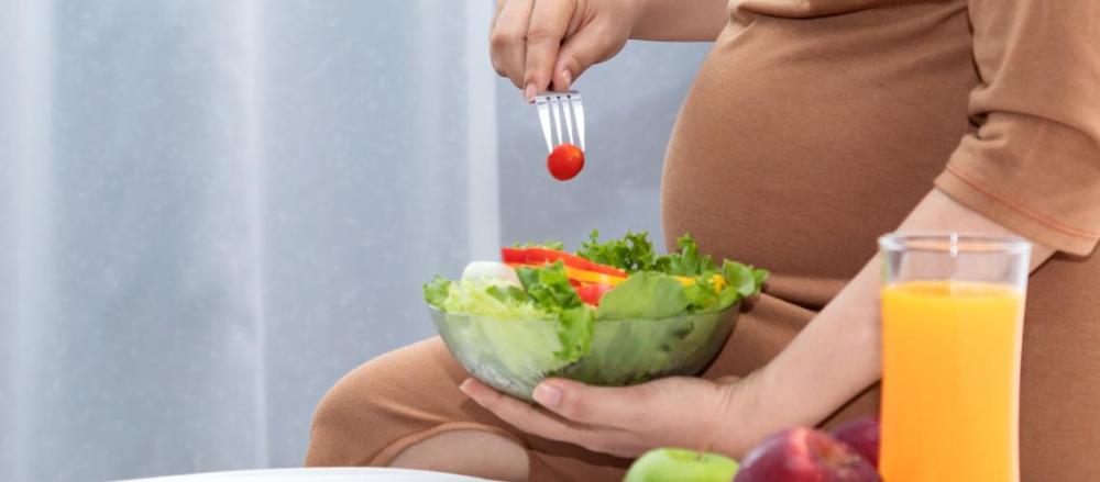 Uma mulher grávida sentada comendo um pote de salada, com um copo de suco ao lado