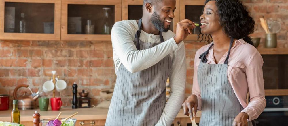 Homem dando pedaço de comida para mulher experimentar, enquanto cozinham juntos