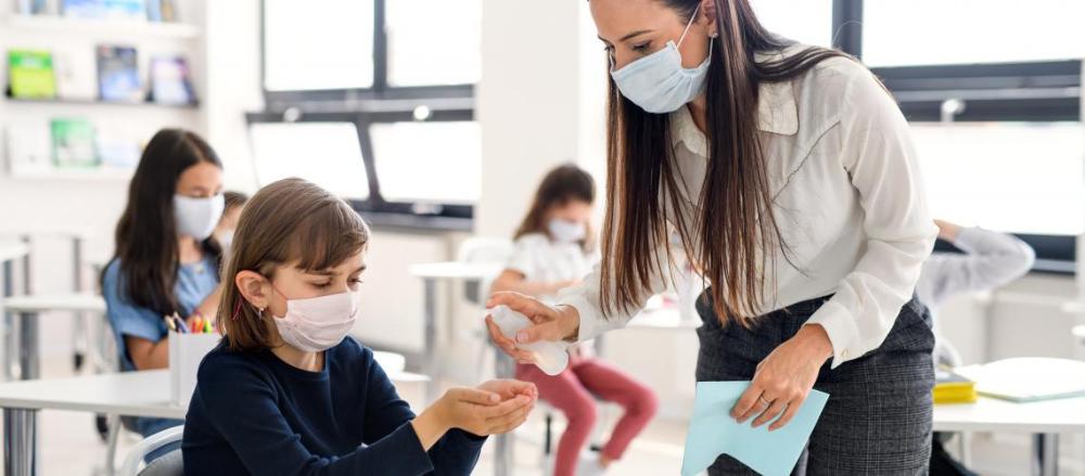 Professora em sala de aula próxima a uma aluna, com todos os presentes usando máscaras cirurgícas