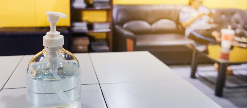 Mesa com um frasco de álcool em gel, com um apessoa sentada em um sofá, de fundo