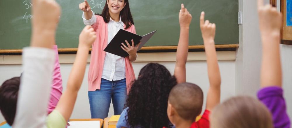 Professora em sala de aula apontando para criança, enquanto todas as outras estão com os dedos indicadores levantados