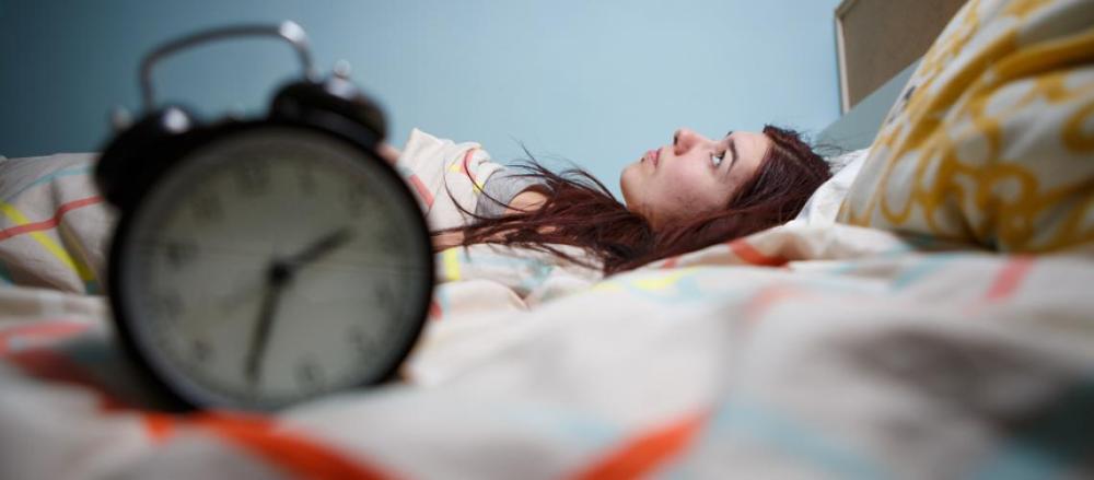 Close de uma relógico, com uma jovem de fundo deitada em um cama, acordada e possivelmente com insônia
