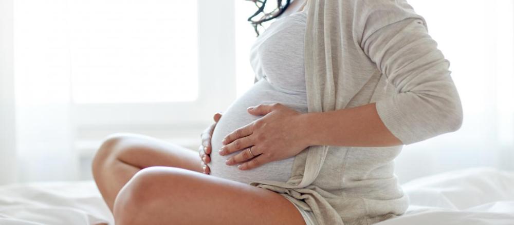 Mulher grávida sentada em uma cama branca, com as mãos na barriga