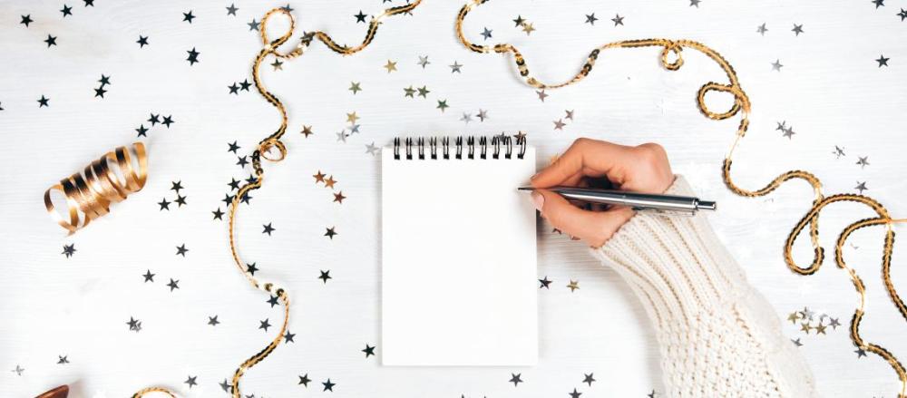 Uma mesa com confetes e estrelas dourtadas e um caderno em branco, com uma mão segurando uma caneta, se prepando apra escrever algo