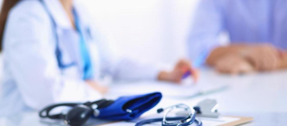 Medidor de pressão e estetoscópio sobre uma mesa, com médico e paciente conversando de fundo