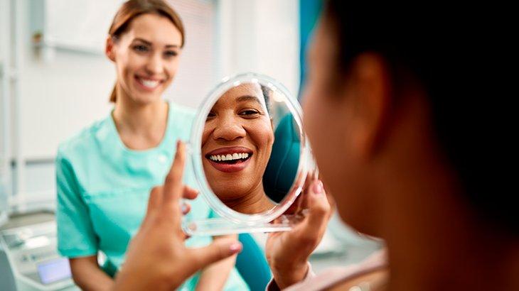 Imagem de uma mulher em um consultório de dentista, sorrindo enquanto se vê no espelho