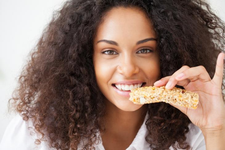 Imagem de uma mulher sorrindo enquanto come uma barra de cereais, indicando como a boa alimentação ajuda a manter a saúde dos dentes