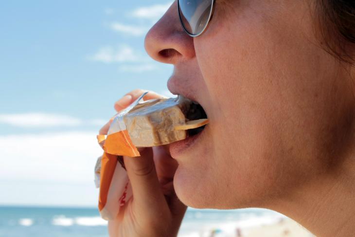 Imagem de uma mulher mordendo um picolé na praia, indicando sensibilidade dental