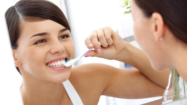 Imagem de uma mulher sorrindo enquanto escova os dentes em frente ao espelho