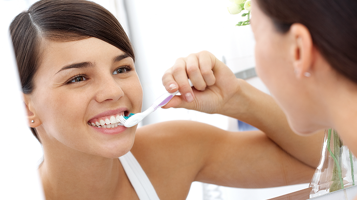 Imagem de uma mulher sorrindo enquanto escova os dentes em frente ao espelho
