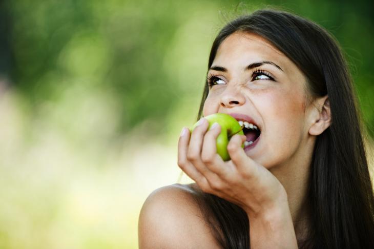 Imagem de uma jovem sorrindo enquanto morde uma maçã verde, destacando a importância dos alimentos para a saúde dos dentes