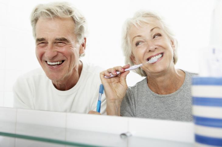 Imagem de um casal de idosos escovando os dentes juntos em frente ao espelho