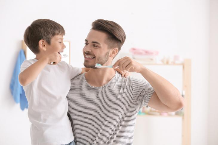 Imagem de um homem com uma escova de dentes nas mãos ensinando uma criança a prática da escovação dental