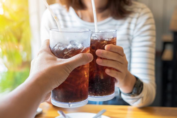 Imagem de duas pessoas brindando com copos de refrigerante, indicando que o consumo excessivo pode prejudicar a saúde bucal