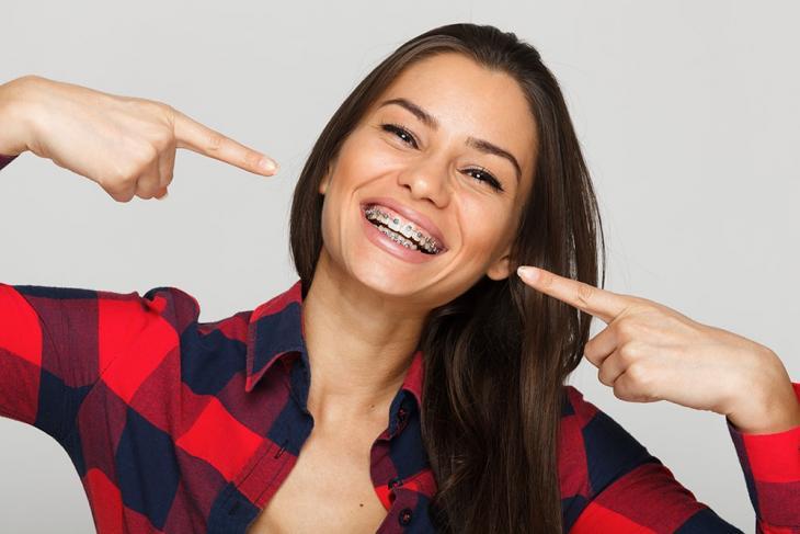 Imagem de uma mulher de aparelho nos dentes, sorrindo, enquanto aponta com ambos os dedos indicadores para a própria boca